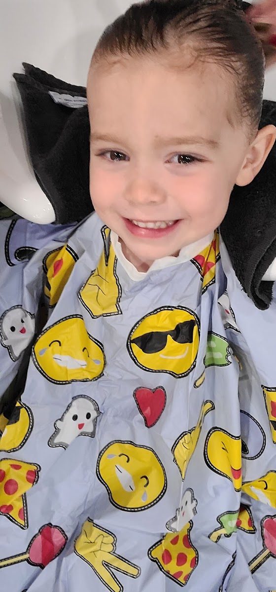 A smiling young client in an emoji-print cape at the shampoo bowl